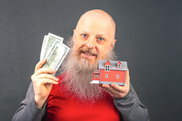 happy senior male with long white beard smiling while holding with cash and a small red house model, representing retirement savings, real estate investment, and wealth management ideas