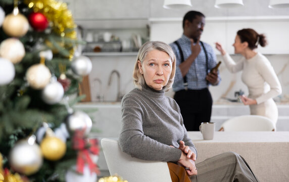 Upset old-aged woman sitting in the kitchen next to Christmas-tree and middle-aged man and woman arguing standing behind