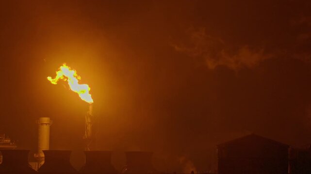 An industrial flare stack burns off excess gas with a large, bright orange flame and thick smoke against a dark night sky. Slow motion footage.