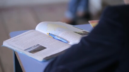 Close up of student's hand pointing at symbols in a textbook. The view shifts to crossed arms resting on a desk near a notebook and a blue pen.

