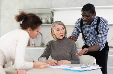 Competitive middle-aged man and woman conflicting and old woman writing testament sitting at kitchen table