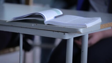 Close up of student's hand pointing at symbols in a textbook. The view shifts to crossed arms resting on a desk near a notebook and a blue pen.
