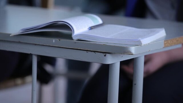Close up of student's hand pointing at symbols in a textbook. The view shifts to crossed arms resting on a desk near a notebook and a blue pen.
