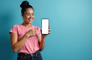 Fototapeta premium Young happy woman laughs, points at mobile phone. Holds modern smartphone with blank white screen. Female recommends new app, online content, tech product to viewers. Shows device for advertising on