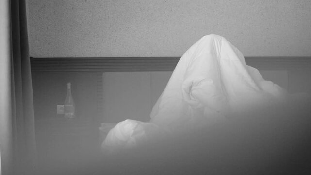 Cinematic black and white shot of restless woman under duvet with long hair covering face. Wine bottle on nightstand suggests severe hangover, headache or morning depression lifestyle.

