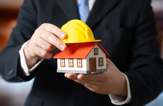 A businessman places a yellow hard hat on a house. The renovation project begins. Construction planning, home repair budgeting and real estate development processes.