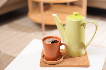 Cup of coffee with coaster and kettle on table in room