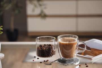 Glass cup of cappuccino with coaster and coffee beans on table in room