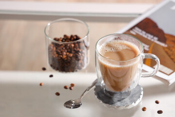 Glass cup of cappuccino with coaster and coffee beans on table in room, closeup