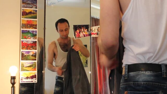 A man stands before a dressing table mirror wearing an undershirt and jeans. He puts on a white dress shirt, getting ready for work or an event.

