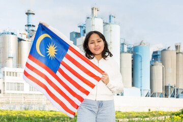 Cheerful young woman standing with big flag of Malaysia against background of factory