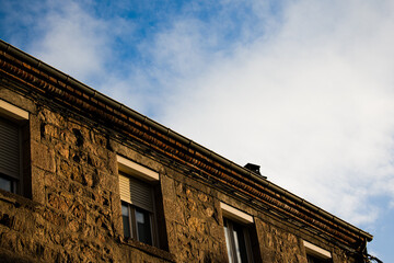 High-contrast city street with deep shadows and bright sky. Dark silhouetted buildings frame a vivid orange facade in the distance. Strong sunlight creates a dramatic, urban atmosphere.