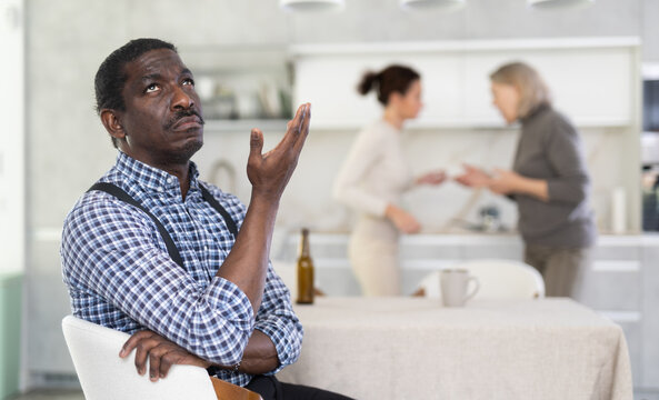 Angry middle-aged man sitting at table while two female family members disputing with each other in the back