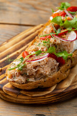 A savory open-faced sandwich adorned with shredded meat, vibrant tomatoes, radishes, and crisp greens rests on a warm wooden board. The cozy kitchen ambiance enhances the appealing presentation