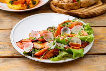 A colorful salad featuring leafy greens, sliced tomatoes, radishes, and herbs is served on a rustic wooden table. The sunlight enhances the freshness of the ingredients