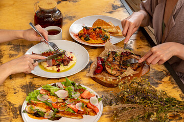 Diners share a delightful meal featuring grilled meats, fresh salads, and rich sauces at a warm, rustic table, showcasing the joy of good food and friendship