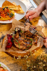 A hand reaches for a succulent grilled chop, garnished with fresh herbs, while a colorful plate of beans and a crisp tortilla lies nearby. Warm tones create a welcoming atmosphere