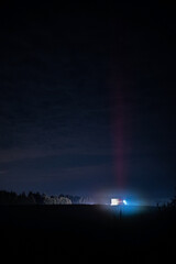 A solitary structure stands in the darkness, illuminated by a bright beam of light cutting through the night. The scene conveys an eerie, otherworldly atmosphere, surrounded by shadows