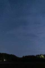 Fototapeta premium Under a vast night sky, stars twinkle above a serene forest. In the distance, a small house glows softly, reflecting tranquility and nature's beauty at night
