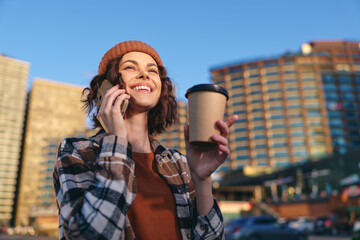 Woman holding coffee and phone in city, smile and beanie in warm sunlight during golden hour;...