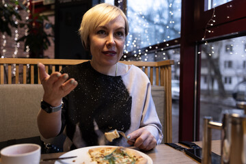 A white woman with blonde hair is sitting at a table, sipping a drink from a white cup. She is wearing a light shawl, and she is surrounded by the soft illumination of a lamp.