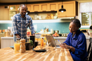 African American young couple making grocery shopping decisions together at home. Focused man and woman checking website to restock household supplies, fresh produce and nonperishable essentials.
