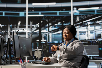 Anxious data center technician checking clock at work, suffering burnout, rushing to finish tasks. Stressed African american woman in server farm checking watch, pressured by deadline