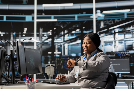 Anxious data center technician checking clock at work, suffering burnout, rushing to finish tasks. Stressed African american woman in server farm checking watch, pressured by deadline