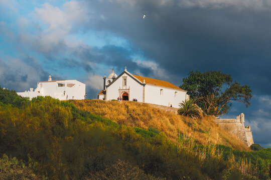 White church stands on a golden hillside under dramatic skies in the village of Cacela Velha, Portugal.