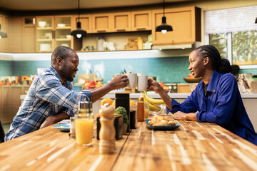 African american couple clinking coffee mugs and enjoying weekend morning together, bonding and relaxing at the breakfast table. Young happy people sharing a positive conversation.