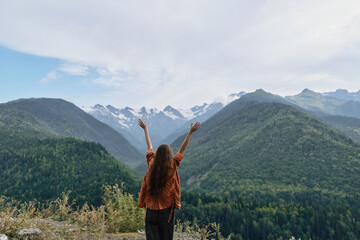 Woman stands on a high viewpoint arms raised embracing nature and mountains a vast green valley under a clear blue sky conveying triumph and freedom during outdoor adventure