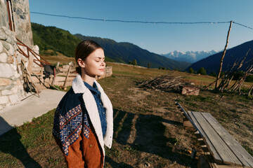 Fototapeta premium Woman portrait in sunlight by rustic cabin with closed eyes, standing in nature overlooking mountains and meadow. Serene outdoor scene with jacket, calm expression and peaceful landscape.