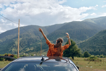 Fototapeta premium Woman stands on a car roof with arms raised enjoying freedom during a road trip in a scenic mountain landscape outdoors, conveying adventure and happiness on the open road