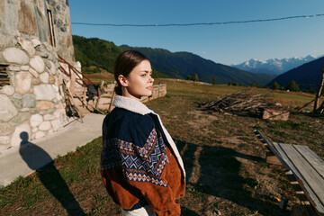 Fototapeta premium Woman portrait in countryside wearing patterned sweater near stone cottage, looking toward distant mountains at golden hour. Outdoor lifestyle, travel, nature and peaceful rural scene.