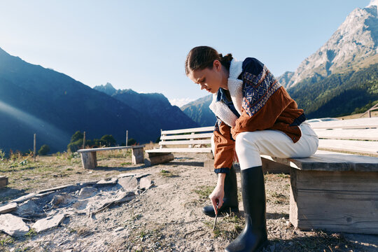 Woman in mountains sits on wooden bench wearing shearling jacket and rubber boots outdoors, adjusting footwear on a sunny alpine trail by rocky ground with panoramic valley view. - Powered by Adobe