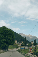 Rural road winds through a countryside landscape with green trees and distant mountains under a bright sky, a small village visible in the background and a calm, open atmosphere.
