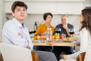 Young guy looking upset and troubled while wife and elderly parents-in-law interacting at table in background during family meal..
