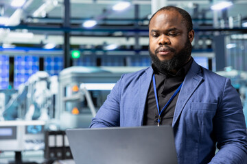 Obraz premium Solar energy black expert in a factory reviews data on laptop, inspecting manufacturing plant performance and symbolizing solar innovation. Industrial employee oversees production line.