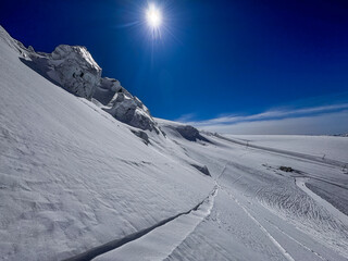 Wide snowy glacier landscape in the Swiss Alps with a bright sunburst, rugged ice formations, and deep winter textures under a clear blue sky.
