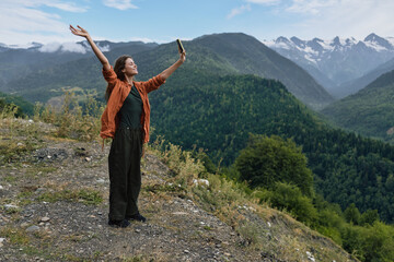 Joyful woman stands on a rocky overlook with arms raised, enjoying panoramic mountains, green valleys, and a clear blue sky in a scenic outdoor setting.