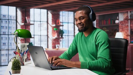 Smiling person in modern loft wearing headphones and listening to music during relaxation time, feeling pleased. Happy guy having fun at home with exciting songs through audio headset. Camera B.