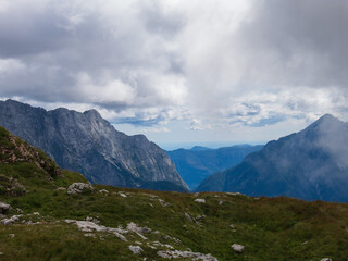 Naklejka premium A panoramic alpine landscape of Montgart in the Alps featuring rugged rocky cliffs, soft green slopes, a winding mountain road and a dramatic cloudy sky