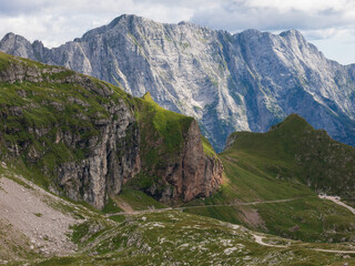 A panoramic alpine landscape of Montgart in the Alps featuring rugged rocky cliffs, soft green slopes, a winding mountain road and a dramatic cloudy sky