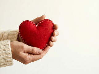 Fototapeta premium Hands gently cradle a vibrant red felt heart, symbolizing love, care, and compassion on a clean background.