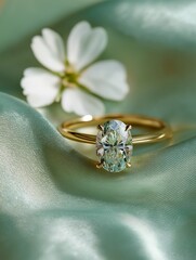 Golden engagement ring with oval diamond resting on soft green satin fabric with white flower in background, gentle natural light creating romantic mood, concept of love elegance and bridal luxury