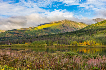 Obraz premium ranquil lakeside scene in Alaska s Chugach Mountains along the Seward Highway (AK-1) toward Seward, with mirrored reflections of golden birch trees and distant mist-veiled peaks in calm waters