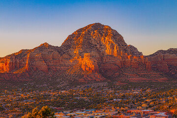 Naklejka premium A warm sunset illuminates the iconic red rock formations of Sedona, Arizona. The glowing cliffs rise above the town below, creating a striking desert mountain landscape.