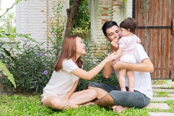 Happy interracial family bonding in garden backyard. Middle Eastern father holding baby while Asian mother playfully touches nose. Parents sitting on grass outdoors.