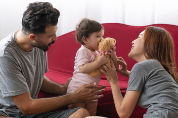 Happy interracial family moments as Middle Eastern father and Asian mother play with cute baby girl holding teddy bear near red sofa. Loving parents bonding with child at home.