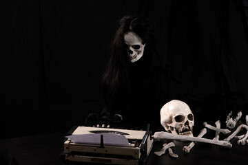 Spooky skeleton ghost with long black hair typing on vintage typewriter in dark room. Creepy grim reaper writing horror story with human skull and bones on table for Halloween concept.